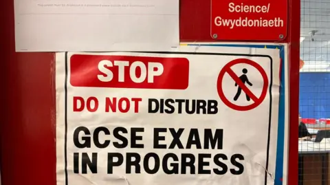 A paper sign pinned to a red door. The door has a sign saying Science and a small window which shows pupils at desks inside the room. The sign reads: "STOP. DO NOT DISTURB. GCSE EXAM IN PROGRESS". The sign is white with red and black writing and also has a stick figure of a person walking with a red cross through it.
