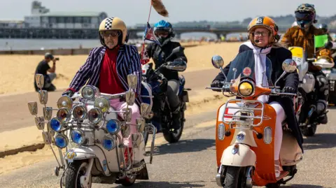 John Coker A man and a woman riding scooters next to one another by a beach. His has lots of lights and mirrors on the front, hers is orange.