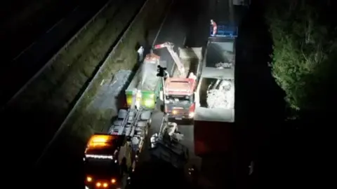 A night-time clean-up operation on a dual carriageway, viewed from an elevated position. A piece of heavy machinery with a lifting arm is positioned alongside the trailer and appears to be removing or handling material from inside it.