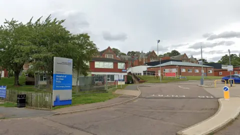 Google The front of the Hospital of St Cross with a blue and silver NHS sign and brick buildings in the background