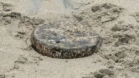 A flat, circular piece of metal can be seen partly buried in the sand. It's rusted, with patches of brown and black and silver flecks underneath.