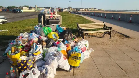 Paul Granger A council bin on a seafront promenade is overflowing with rubbish and dozens of carrier bags, also full of empty cans and bottles, and piled up next to it.