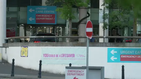 Taken outside a hospital the picture shows blue signs with 'urgent care centre' and 'emergency department' written on them. There is a white 'you could be a life saver sign' as well on the white wall in front of the hospital. The hospital itself has a glass front.