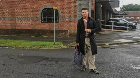 Ian Trowell A man stands on a wet street outside a brick building, holding a shopping bag and wearing a long dark coat. The surroundings include parked cars, a sidewalk, and a small patch of grass near the building. The overcast sky and damp road suggest recent rain, giving the scene a muted, everyday atmosphere.