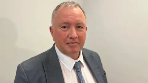 BBC Deputy Liam McKenna - A man with black and grey hair looking towards the camera. He's wearing a blue suit jacket, a white shirt and a blue and white dotted tie. 