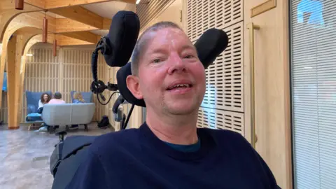 A man wearing a blue jumper is smiling at the camera. He is sat down in a wheelchair.