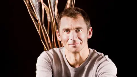 Brendon Murless A man smiles softly at the camera on a black backdrop