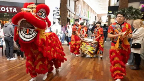 Solihull Council Two performers in a Chinese dragon costume and four other men in bright red and orange and orange decorated festival costumes walk through the centre of a shopping centre, watched by crowds. Three of the men are carrying a large drum, wrapped in a yellow-patterned cloth.