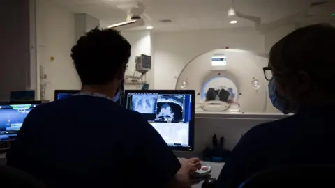 Medical staff at a hospital look at a computer screen showing images while a patient is undergoing a CT scan exam in the neighbouring room.