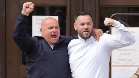 Two men with their arms around each others shoulders with their hands raised smile outside a court building