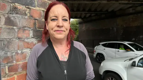 Tracy looks into the camera while leaning against the brick wall of Fisherton Bridge. Two white cars are in the background. She has red hair and is wearing a zip black top.