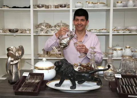Eddisons A dark haired man wearing a lilac shirt is standing behind a wooden table. He is pouring water from a silver claret jug into a gold-rimmed glass. On the table are a soup tureen, a black lion sculpture with red eyes, canteens of cutlery and crystal decanters.
