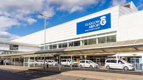 The exterior view of a large airport building. A row of taxis are parked outside it with a large sign - with whitter lettering on a blue background reads Glasgow Airport - proud to serve Scotland.