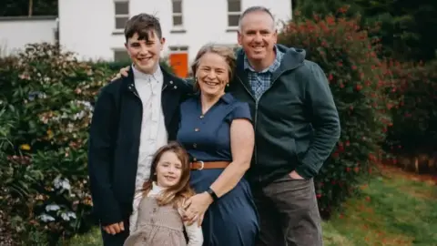 A family photo with a woman, a man a little girl and a teenage boy. They are outside. The mum and daughter are wearing dresses. The dad and teenager have jackets on. Bushes are behind them. 