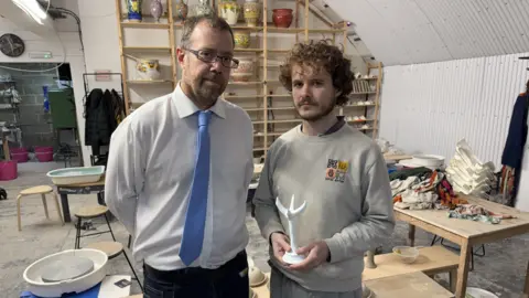 Simon Balch Simon Balch smiles at the camera while standing next to another man who holds an egg cup in the shape of a chicken's foot. Simon wears a white shirt and blue tie, while the other man has short curly hair and wears a grey jumper.