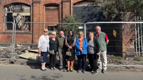 A group of seven people, with a mix of men and women, stand in front of a derelict building on a sunny day. There is grey fencing around the building.