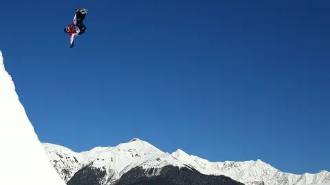 Getty Images Aimee Fuller in a red and white coat mid jump with blue sky and mountain tops in the background 