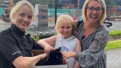 Lancashire Police PC Liz Harrison holding a box containing the black cat, with Caroline and her daughter smiling and stroking the cat