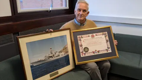Northumberland County Council Glen Sanderson, with short white hair, wearing a beige jumper over a blue shirt and grey pants, sits on a sofa. He is holding up two frames, one shows a large navy ship and the other shows a certificate showing the Freedom of Northumberland registration. 