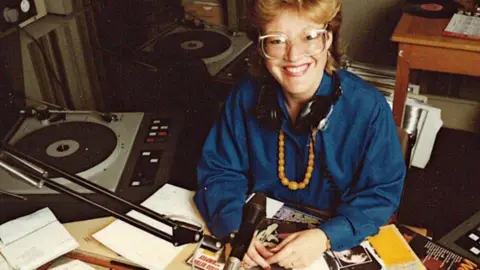 A woman sitting at a radio desk with a microphone and turntable and magazines over the desk. She is wearing a blue shirt and headphones round her neck and smiling up at the camera