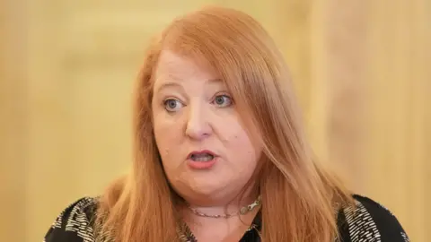 Ms Long speaking as she looks to her left, she has long ginger hair, pink lipstick and is wearing a black and white patterned top with a silver chain necklace.