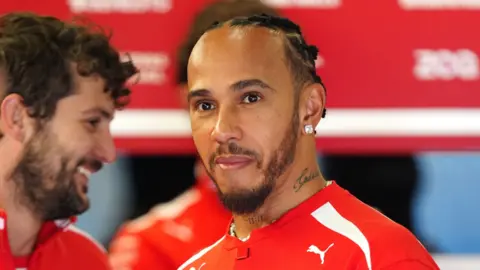 PA Media Lewis Hamilton is wearing a red and white top, has short black hair in tight cornrows and has a trimmed black beard. He is starting to smile. 