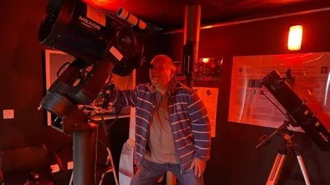 Poltimore Observatory Publican Alan Boddington is wearing a blue and white striped jacket and looking through a very large telescope. There is a smaller telescope to his right.