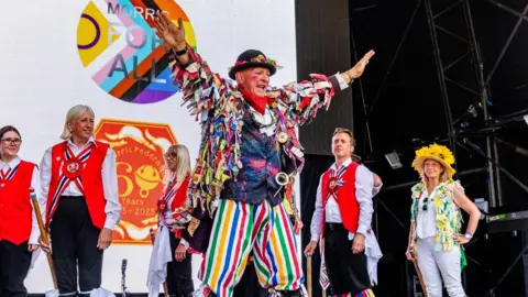 Camp Bestival A group of Morris dancers are pictured performing on stage. At the head of the group, a man in rainbow-coloured clothing and a bowler hat dressed in flowers extends his arms to the sides.
