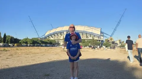 Kirsty Cummings Kirsty Cummings is standing outside a large stadium with her arms around her daughter. They are both in blue, Sunderland's away strip. 