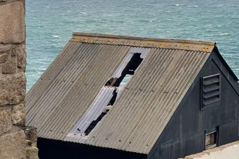 National Trust A tin roof with a large hole in it. In the background is the sea.