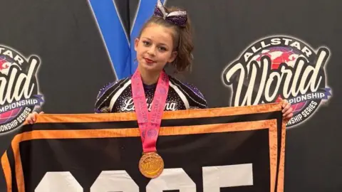 Swindon Lightning Cheerleading Club Abbie Lainton with long brown hair tied in a pony tail wearing her purple and black Swindon Lightning Cheerleading Club kit and a large bronze medal around her neck, holding an All Star World Championship 2025 towel - with a black background.