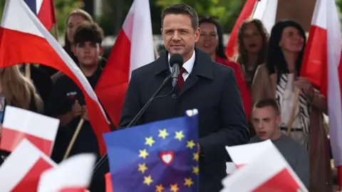 Anadolu via Getty Images Wearing a dark coat and tie, Rafal Trzaskowski, is surrounded by red and white flags