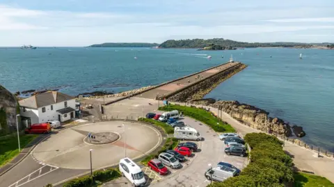 An aerial view of the Mountbatten car park in Plymouth with cars and vans in the car park and the breakwater stretching out into the sea.