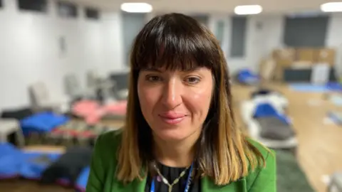 Izzy has long straight hair and is wearing a green jacket 