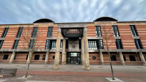 A large three storey court building made from red bricks, with long narrow dark windows and a pyramid shaped porch roof supported by four large stone columns around the main door