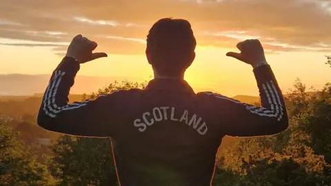 A man wearing a top which says SCOTLAND on the back, with his back to the camera, and a sunrise in the distance.