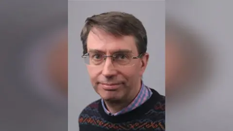 SODC David Rouane headshot in front of a grey background. He has short brown hair, glasses and is wearing a colourful knitted jumper.