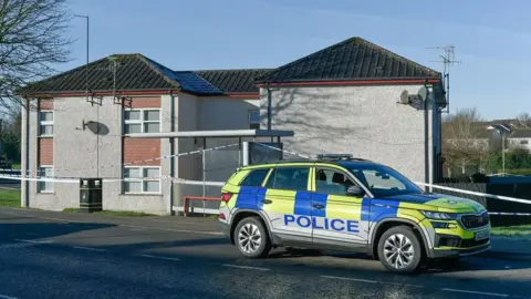 PACEMAKER A white police cord surrounds a block of floors in SELLOINEY Park on the Lisnahull estate in Dungannon. It is a U - -shaped two -story building with gray rendering and pink tile panels that surround their white windows. A blue and yellow police vehicle is parked in front of the cordon.