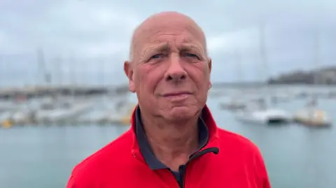 A bald man wearing a red jacket looking towards the camera in front of St Peter Port's Harbour.