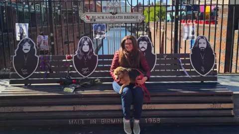 A person sits on a wooden bench holding a small dog on a red leash in front of a canal-side walkway. Behind the bench is a black iron gate with a sign reading “Black Sabbath Bridge,” along with posters and life-size cutout silhouettes of band members. The bench has engraved names “Geezer,” “Ozzy,” “Tony,” and “Bill,” and the text “Made in Birmingham 1968.” In the background, a red-brick building with a clock tower rises to the left, while modern buildings and a canal stretch into the distance under a bright blue sky with scattered clouds.
