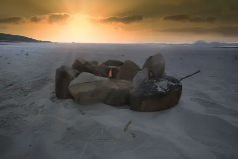 Garry Lynch Stones with a small fire in the middle on a sandy beach at sunset
