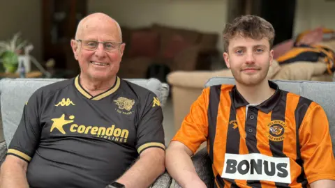 Geoff Golliker is sitting next to his grandson Jacob Allmendinger. Golliker has a bald head and is wearing glasses and a black Hull City T-shirt with yellow branding on. Allmendinger has short brown hair and is wearing an orange and black Hull City football shirt. They are both sitting on grey chairs smiling at the camera. 