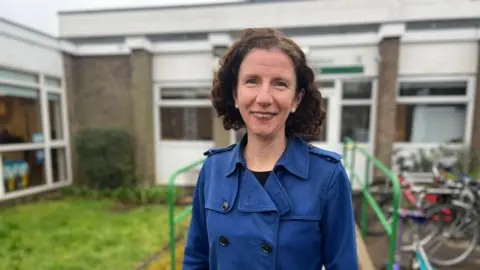 BBC Anneliese Dodds stands in front of a school in a blue coat. She has dark hair, almost at shoulder length.
