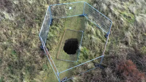 A hole in the ground, from above, surrounded by six metal fences. The hole is in the middle of a grassy area which is surrounded by longer rough grass like the type found in sand dunes. A fence panel is resting on top of the hole that is in the middle of the image.