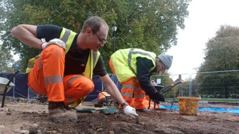 University of Leicester Archaeological Services A man and a woman in hi-vis sift through the dirt for historic artefacts