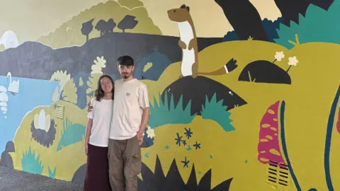 Vinny Lynch and Gemma Pasmore are standing in front of the mural smiling at the camera. He has his arm around her shoulders. He has black hair and a black beard and she has shoulder length brown hair. They are both wearing white T-shirts and she is wearing a skirt and he is wearing cargo trousers. The mural shows a stoat standing up on its back legs which is two thirds the height of the students. There is a swan on the lake and on the shore a nest with three large eggs in it.