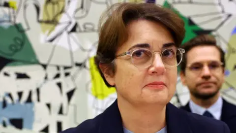 Head and shoulders picture of Laurence des Cars wearing glasses and standing in front of an artwork which is part visible behind her