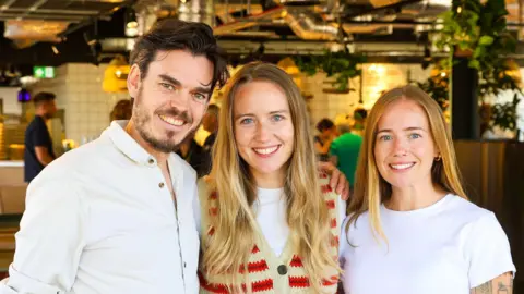 Louis Alder Day Release's three co-founders Paul Humpherson, Amy Taylor and Tess Taylor smiling for a picture inside. Staff and customers are seen behind them.