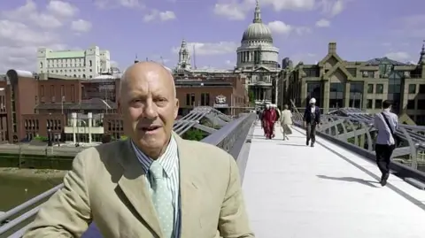 John Stillwell/PA Media A man wearing a light coloured jacket, tie and striped shirt standing on the Millennium Bridge in London. People are walking across the footbridge behind him. St Paul's Cathedral is in the background.