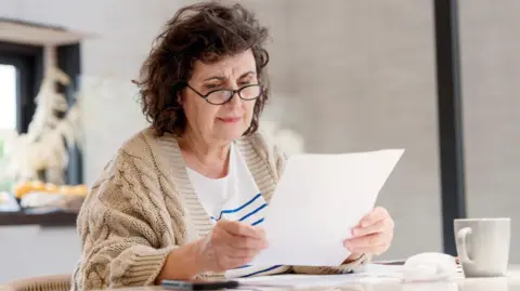 Getty Images An older woman wearing glasses sits at a table looking at a piece of paper.
